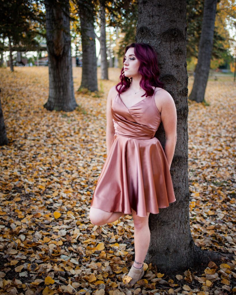 Teenage girl with purple hair in pink grad dress leaning against a tree.